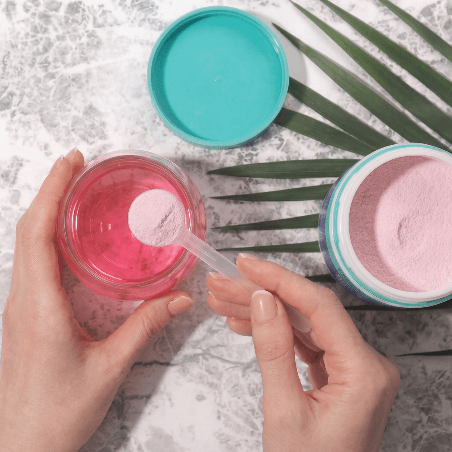 Person holding a jar of pink powder with a spoon, surrounded by palm leaves on a light background